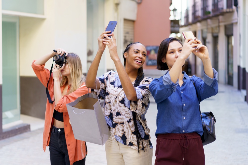 Group of glad diverse female friends taking pictures of street with smartphones and photo camera while standing on sidewalk near buildings. Content multiracial friends taking photos of street