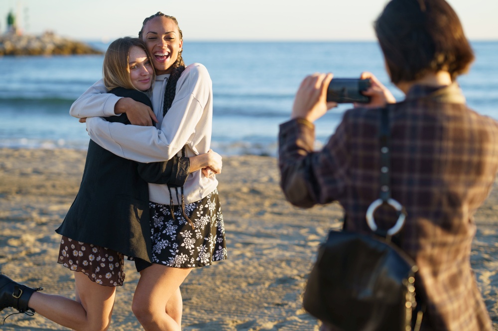 Unrecognizable female photographing cheerful multiracial female friends embracing each other while standing on sandy beach near rippling sea on sunny day. Faceless woman taking photo of hugging diverse girlfriends on seashore