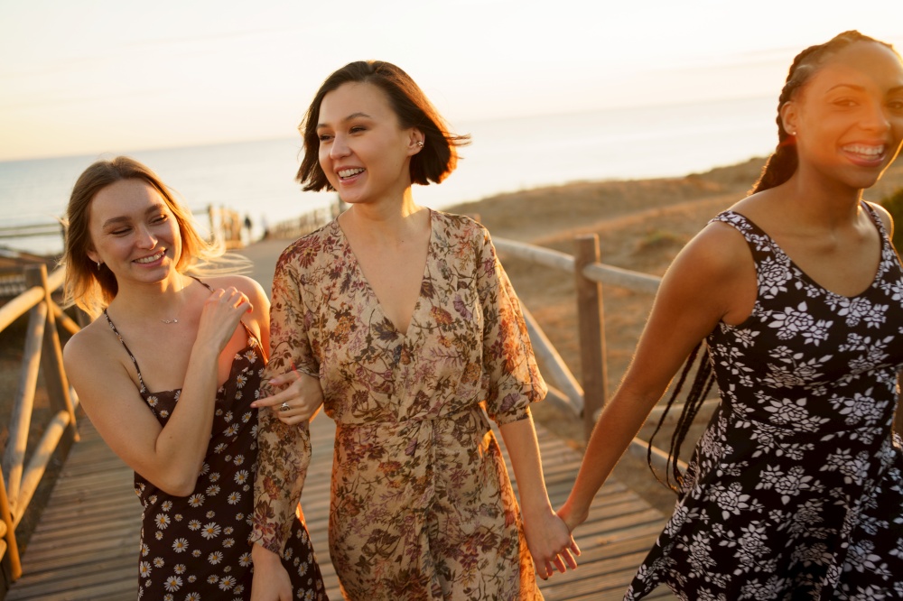 Optimistic multiracial female friends holding hands and walking together on wooden boardwalk while spending time together on beach against sea. Content multiethnic girlfriends walking on footbridge on seashore