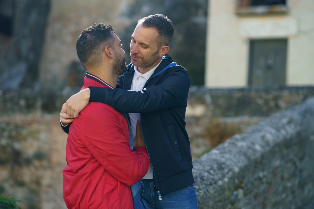 Cheerful loving couple of homosexual males hugging in street and looking at each other. Couple of gay men embracing in city