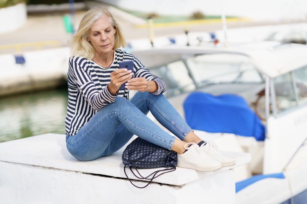Mature woman using her smartphone sitting in a sea port. Elderly female enjoying her retirement at a seaside retreat.. Mature woman using her smartphone sitting in a sea port.