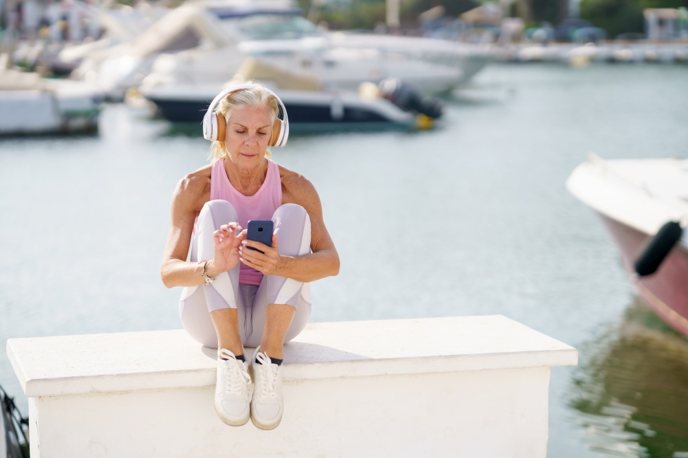 Elderly female athlete in activewear and headphones sitting on embankment in port and listening to music while browsing smartphone. Senior sportswoman listening to music during training