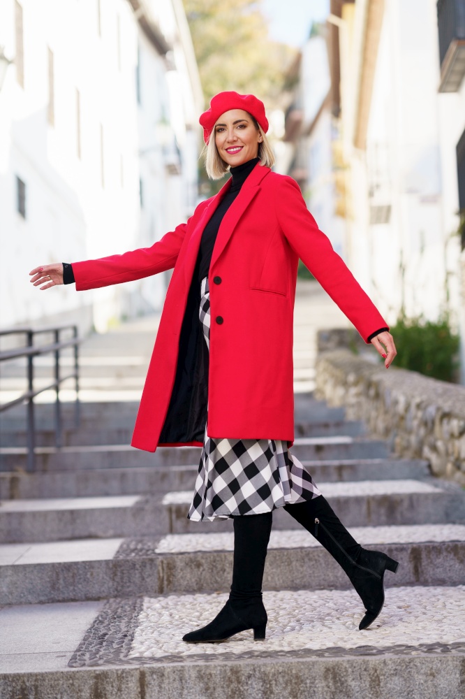 Happy blond woman in trendy coat with dress and beret with boots looking at camera while walking on street steps. Lady in trendy outfit on street stairs
