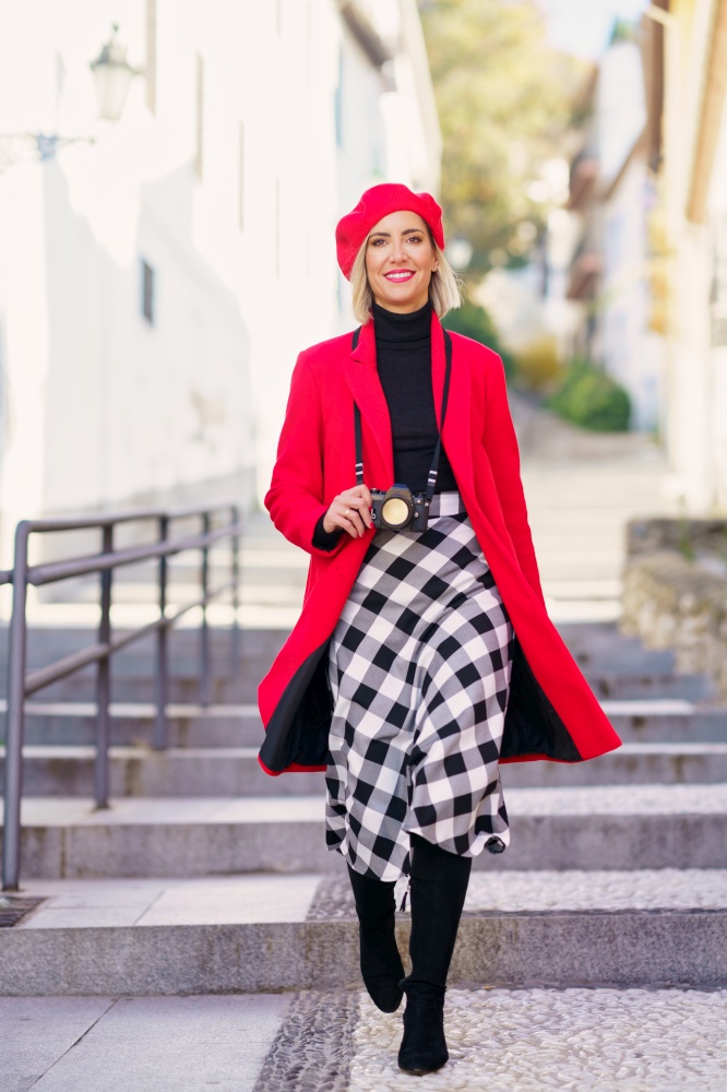 Full length of self assured young lady, with blond hair in elegant outfit and beret, smiling and looking at camera while walking on stone stones with photo camera. Smiling fashionable woman walking on street with photo camera