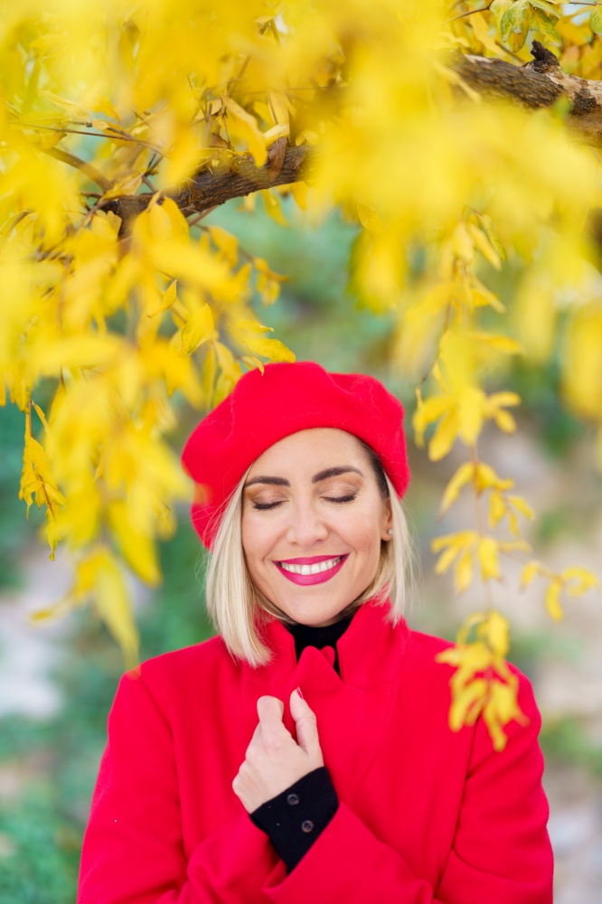 Optimistic young stylish female with blond hair and makeup, wearing red beret and coat, while standing under bright yellow tree in park on sunny autumn day. Delighted elegant lady smiling and enjoying autumn nature with closed eyes