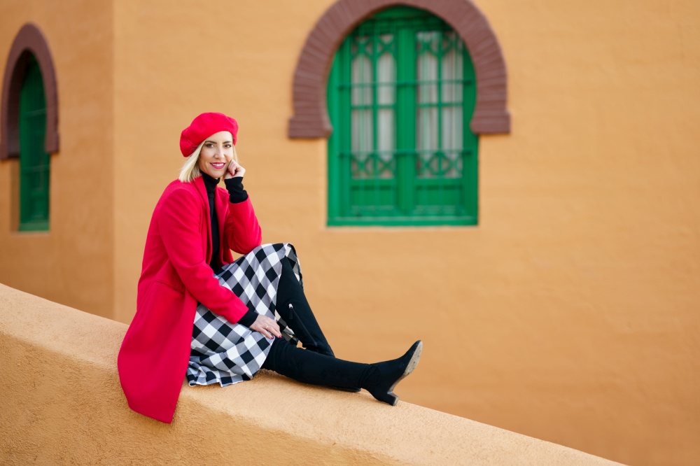 Side view full body of happy young female, in stylish red coat and beret, leaning on hand and smiling at camera while sitting on stone border near aged typical building. Smiling graceful lady relaxing on old town street during vacation
