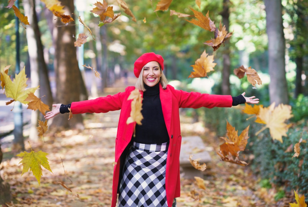 Happy female wearing red beret and coat tossing yellow dry leaves on autumn street and looking at camera. Cheerful woman throwing maple leaves in air