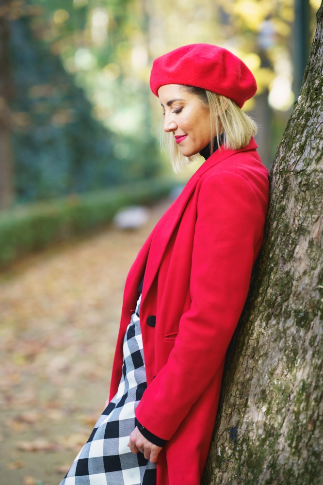 Side view of female wearing red beret and coat with checkered skirt standing near tree trunk in autumn park and looking down. Gentle woman leaning on tree in autumn park