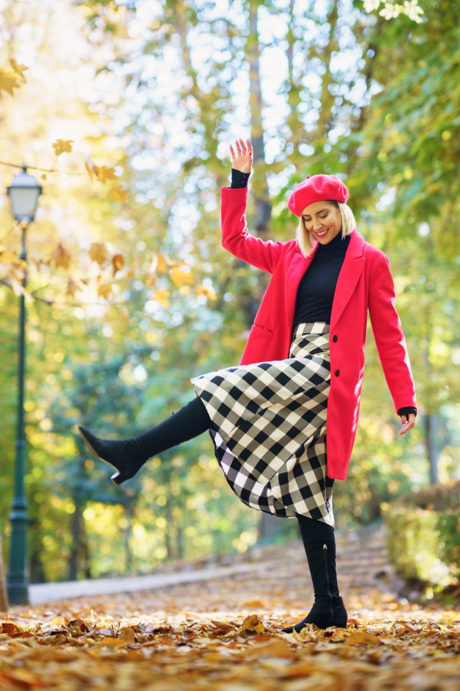 Full body of positive female in red coat and beret raising leg while standing on pathway with fallen leaves in park. Content woman in elegant wear raising leg in autumn park