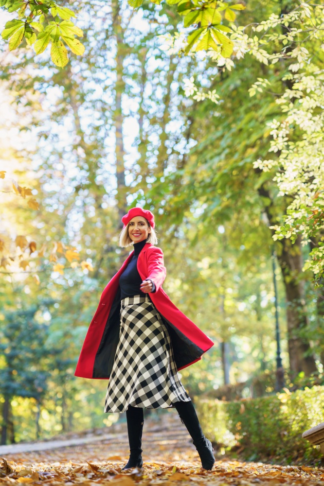 Full body of active female in red coat and beret looking at camera while standing on pathway on autumn day. Smiling woman in autumn park