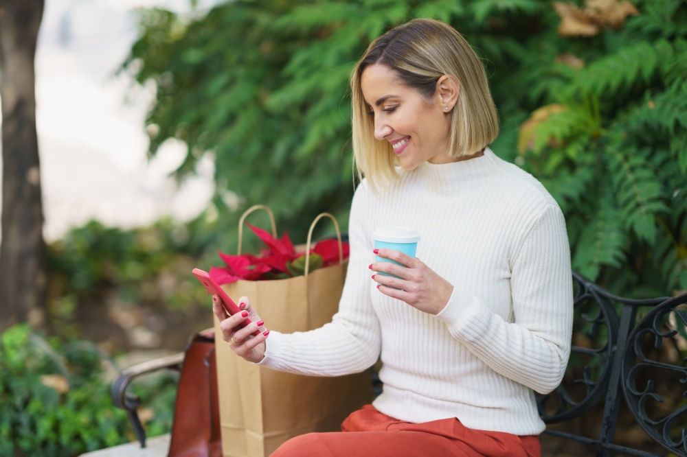 Positive female sitting on bench near paper bag with houseplant and handbag and using cellphone while having takeaway drink on bench in park. Smiling woman using smartphone and drinking beverage on bench
