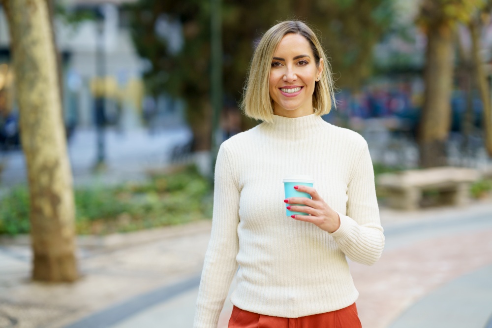 Positive female in white turtleneck with cup of takeaway beverage standing in city park and looking at camera with toothy smile. Smiling woman with cup of drink standing on street