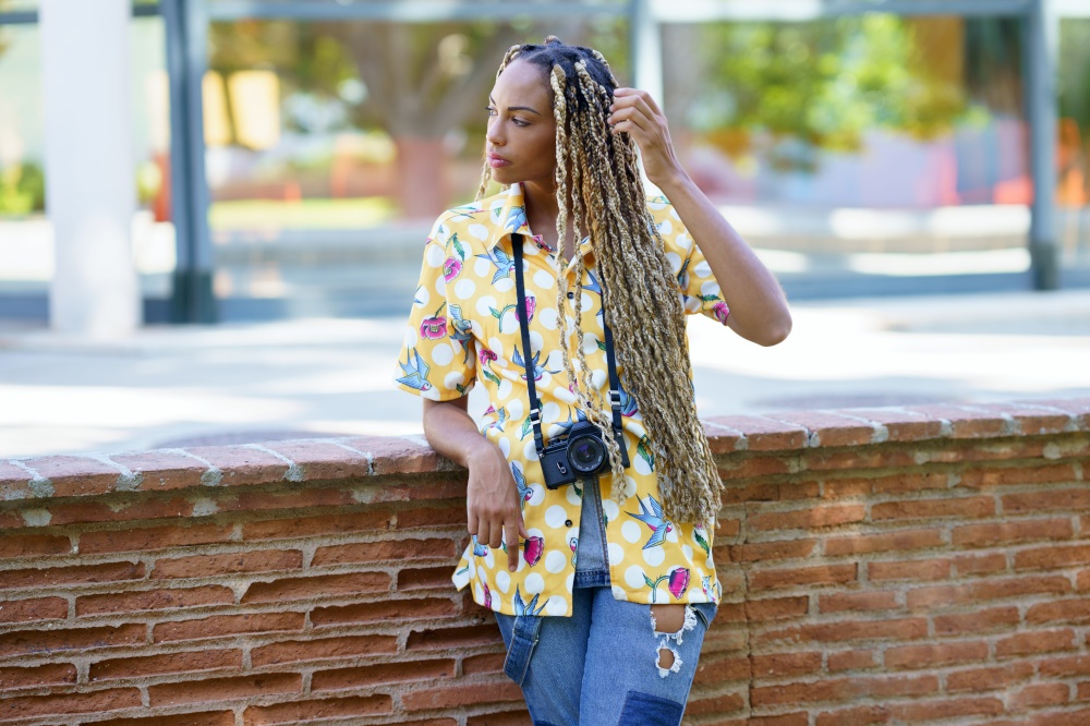 Beautiful African influencer combed with colorful braids standing with a camera outdoors.. African influencer combed with colorful braids standing with a camera outdoors.