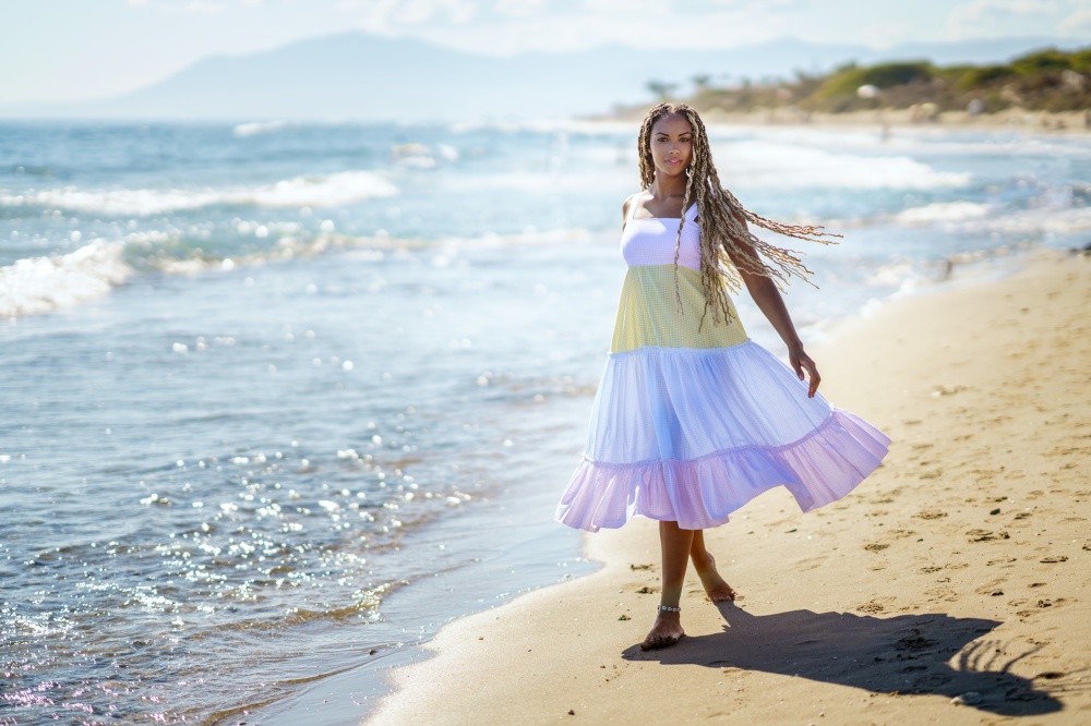 Full body barefoot African American female in summer dress with long braided hair walking on sand near waving sea on sunny day on resort. Black woman walking on beach near sea