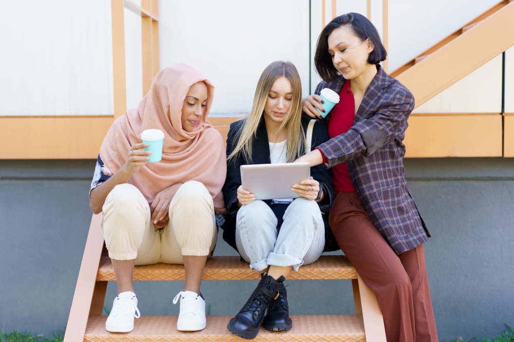 Trendy young multiracial girls students in casual clothes drinking takeaway coffee and using tablet while sitting on stairs in city and preparing for exams. Stylish diverse women friends sharing tablet and drinking takeaway coffee on street