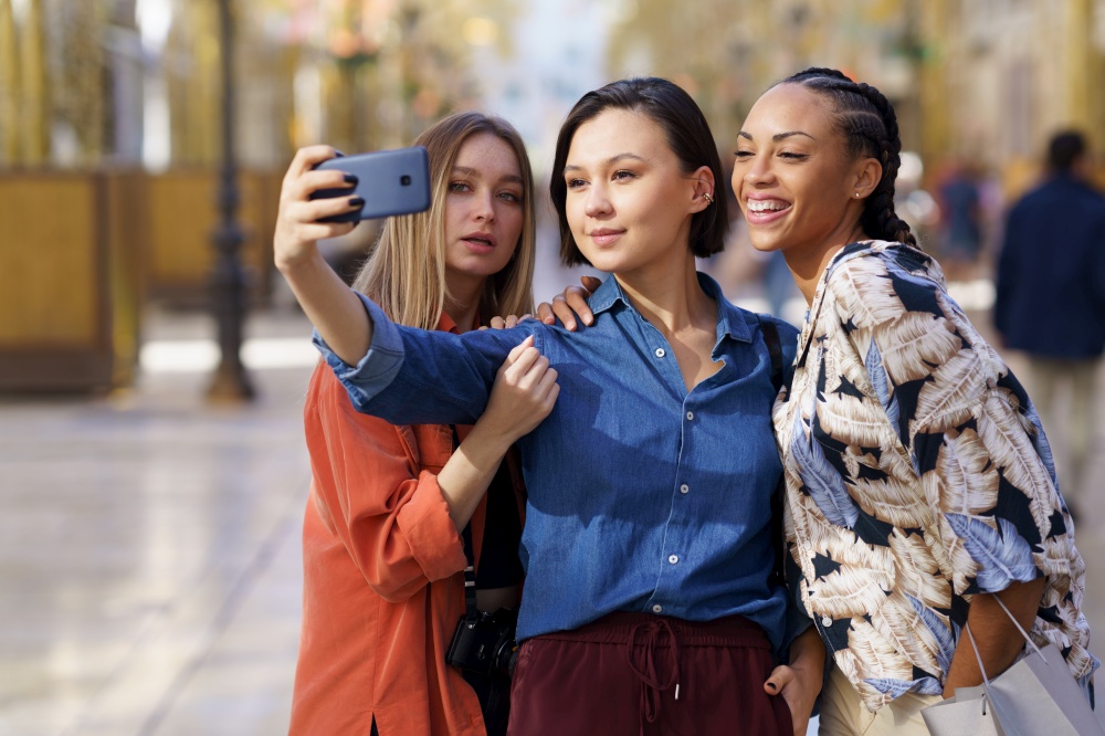 Content multiracial female friends taking self portrait on modern cellphone while standing together on sidewalk against blurred background of city. Trendy girlfriends taking selfie on street
