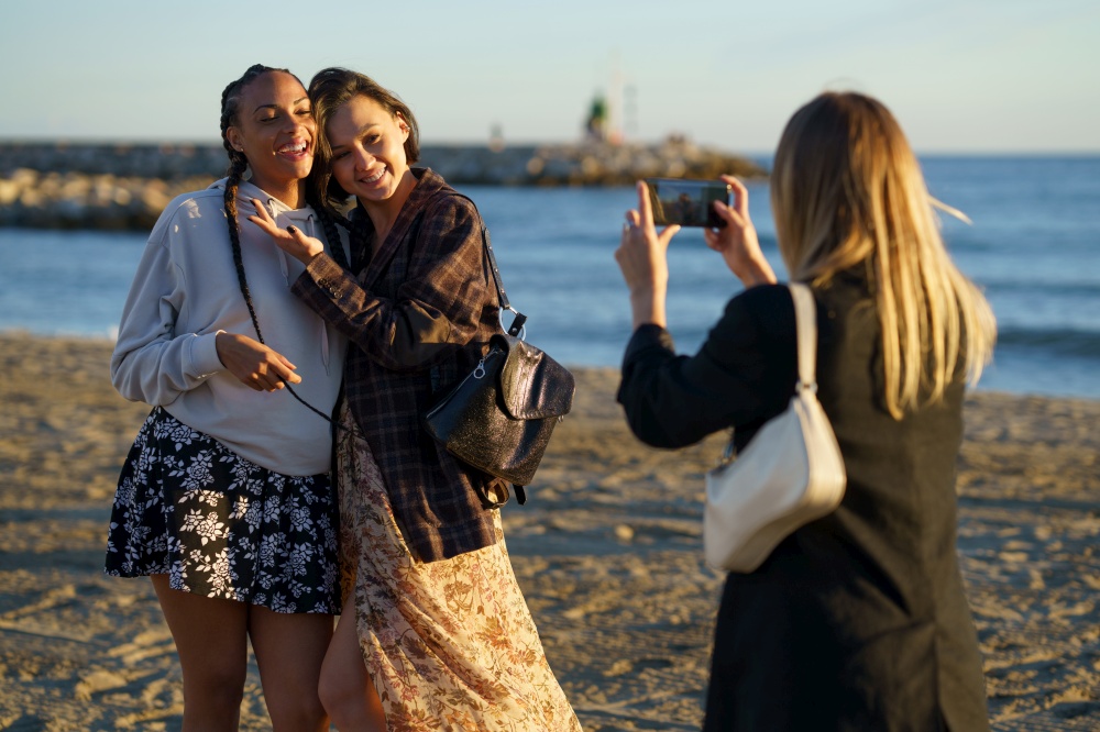 Anonymous woman taking photo of positive multiracial female friends hugging while standing on sandy coast near rippling sea on sunny day. Faceless woman photographing cheerful diverse girlfriends on seashore