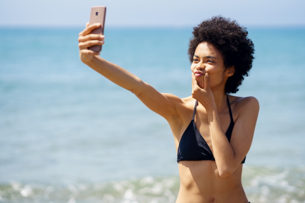 Black woman with afro hairstyle taking a selfie with a funny gesture on a tropical beach. Black girl enjoying her holiday on the beach.. Black woman with afro hairstyle taking a selfie with a funny gesture on a tropical beach.