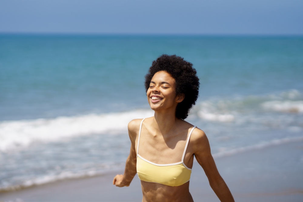 Carefree young African American female tourist, with dark curly hair in stylish swimwear, smiling happily with closed eyes while enjoying sunny summer day on sandy beach. Happy black fit woman smiling with closed eyes on sandy seashore