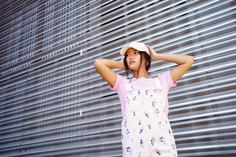 Low angle of glad Asian female in casual clothes, wearing cap, looking away standing near modern building.. Low angle of glad Asian female in casual clothes, wearing cap, standing near modern building.