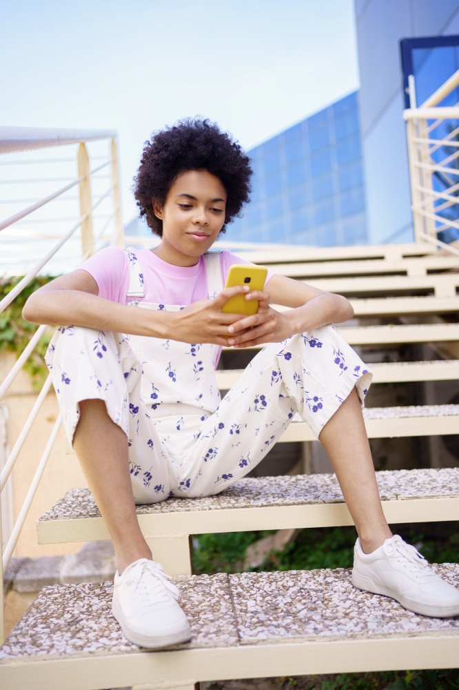 Black woman texting with her smartphone, sitting on steps of a modern building. Cheerful young female, afro hair, on urban background.. Black woman texting with her smartphone, sitting on steps of a modern building.