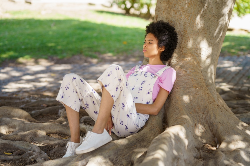 Full body side view of young wistful African American female in casual clothes sitting near thick tree trunk in park. Pensive black woman sitting near tree