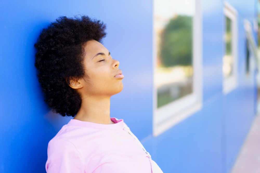 Side view of calm young African American female with Afro hairstyle standing near blue house with closed eyes on street. Black woman standing near building