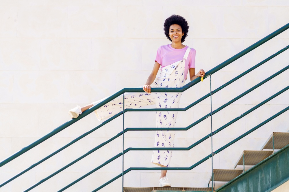 Full body of smiling African American female in casual clothes looking at camera while standing on staircase with leg on railing against white background. Cheerful black woman standing on stairway
