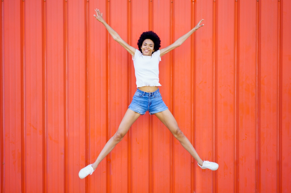 Full body of optimistic African American female in casual clothes looking at camera while star jumping on red background in city. Joyful black woman jumping near wall