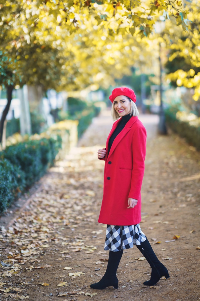 Full body of cheerful female in red clothes looking at camera while standing on pathway in park on autumn day. Stylish woman in an autumn urban park