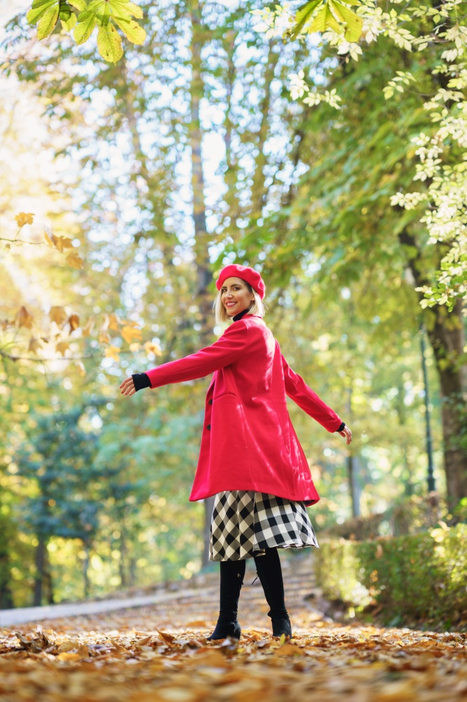 Full body side view of positive female in red clothes spinning around while standing on alley with dried leaves in autumn park. Stylish woman spinning around in park