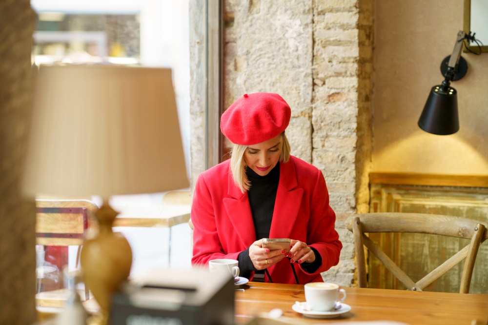 Attractive female in red outfit text messaging on cellphone while sitting at table with cup of coffee in light cafeteria. Elegant woman browsing smartphone in cafe