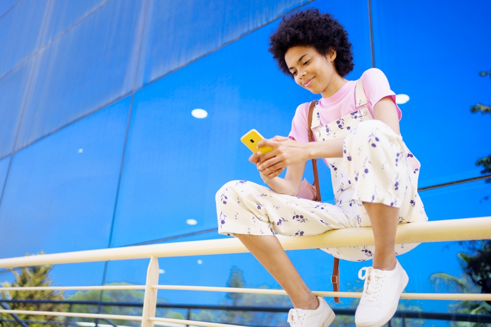 Glad African American female with Afro hairstyle in overall browsing cellphone while sitting on railing near glass building in city. Content black woman texting message on fence