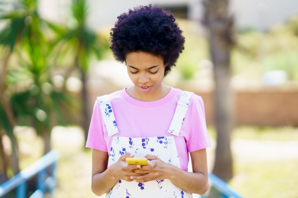 Glad African American female in casual wear scrolling social media on cellphone while standing on street with trees against blurred background. Content black woman checking smartphone on street