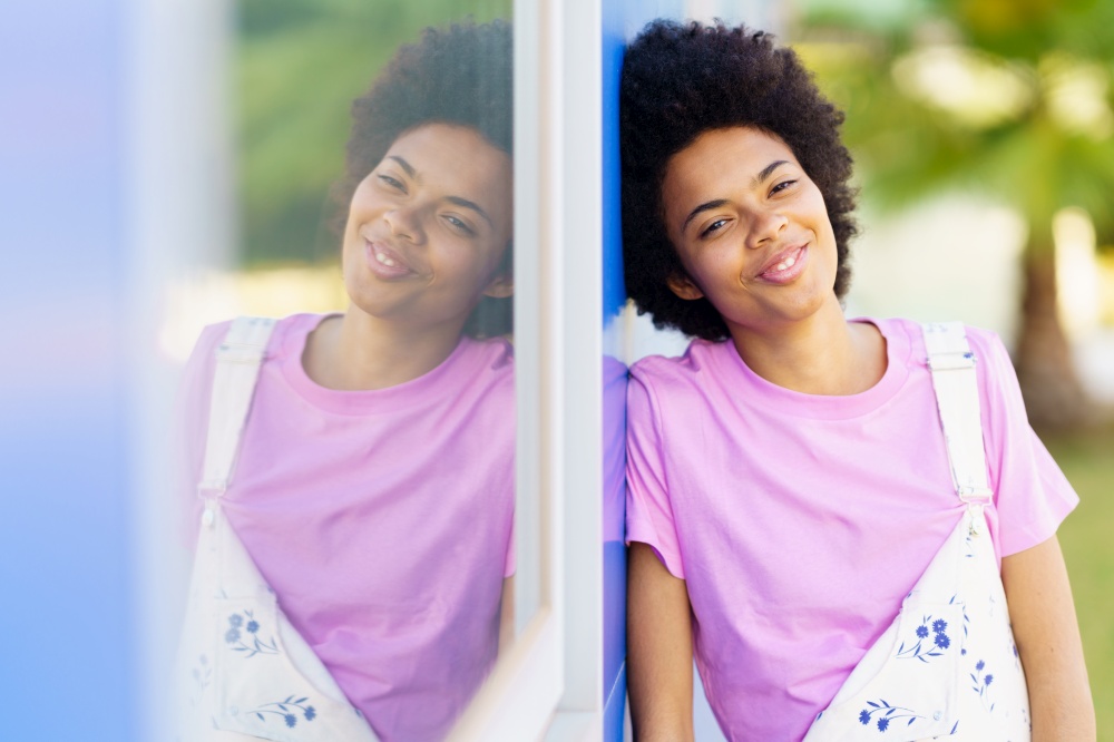 Happy African American female in casual clothes looking at camera while standing near window of house on street against blurred background. Positive black woman standing near building