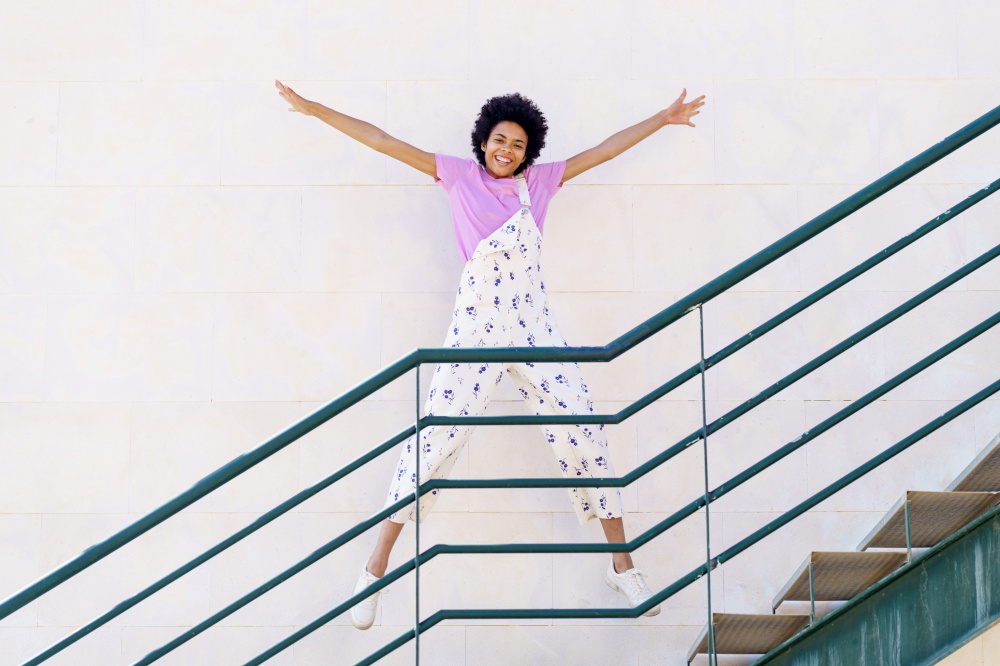 Full body of optimistic African American female in overall looking at camera while star jumping on staircase against white background. Cheerful black woman jumping on stairway