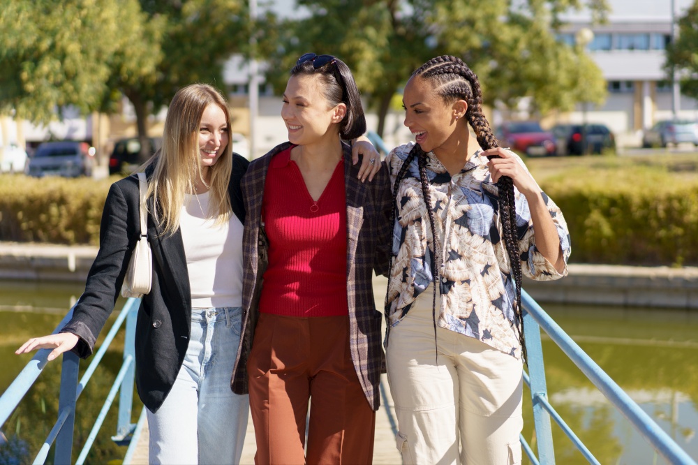 Group of content diverse female friends strolling on footbridge crossing river on sunny summer day in city with green trees. Cheerful multiracial women on footbridge