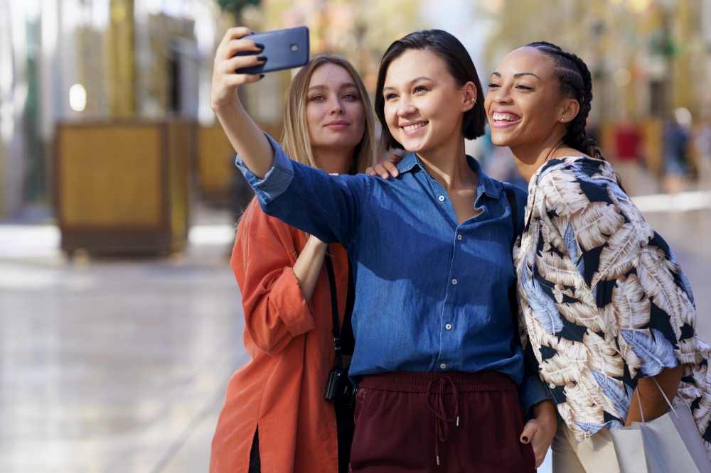 Group of cheerful diverse female friends taking self portrait on cellphone while standing on sidewalk against blurred background in city. Joyful multiethnic women taking selfie on smartphone