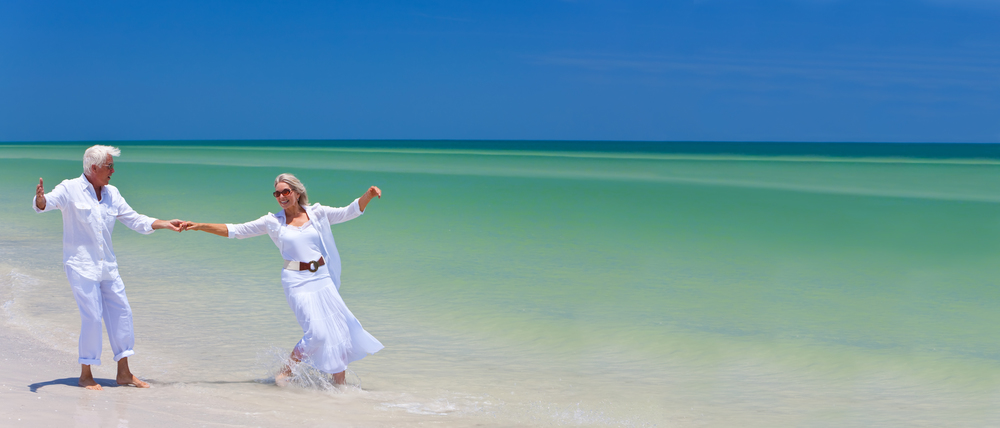 Panoramic web banner happy senior man and woman couple dancing and holding hands on a deserted tropical beach with bright clear blue sky