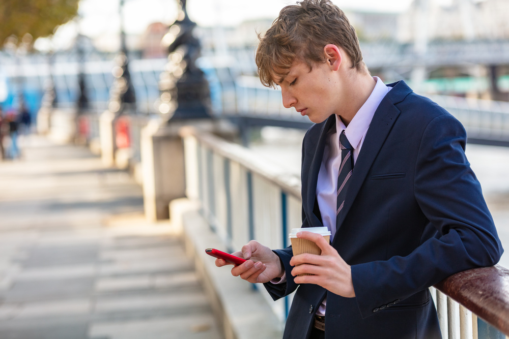 Male young adult teenager wearing suit and tie using smart cell phone for socail media and drinking takeout coffee