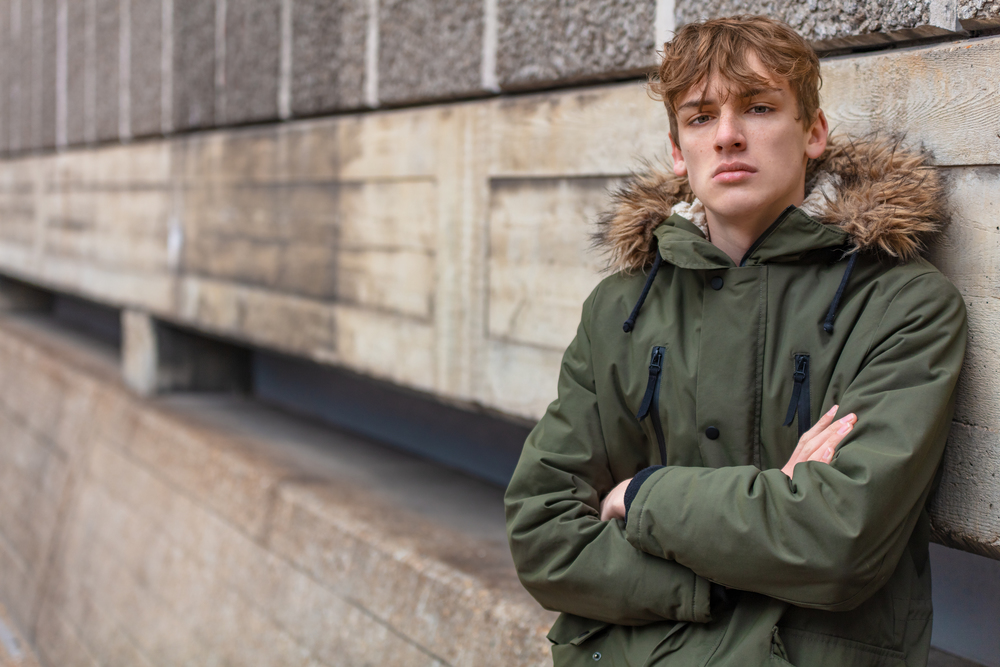 Young adult male teenager boy outside in an urban city wearing a green parka coat leaning against a wall