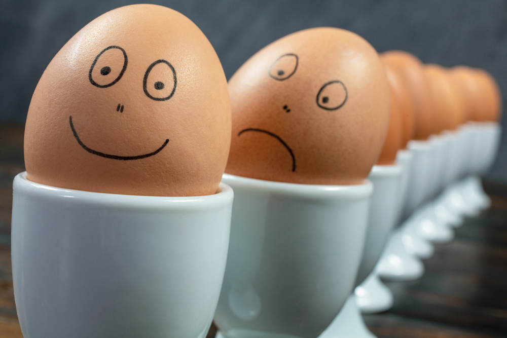 Concept waiting queue or line up of ten boiled eggs in white egg cups with drawn happy and sad faces on a wooden table