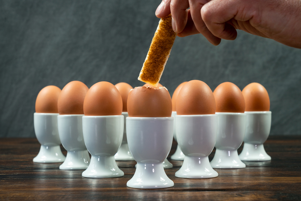 Person dipping toast soldier into one boiled egg of a group of eggs in white egg cups on a wooden table