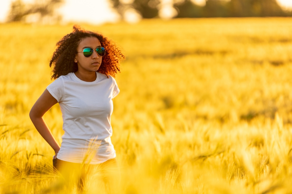 Beautiful mixed race African American female teenager young woman in a field of wheat wearing white t-shirt, sunglasses at sunset