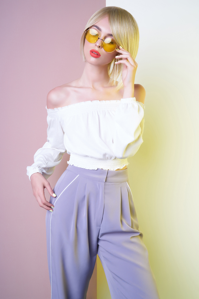 Beautiful sexy blonde with professional classic make-up pose in photography studio. Sensual stylish woman in blue pants. Blue-eyed lady with perfect lips in modern colour sunglasses