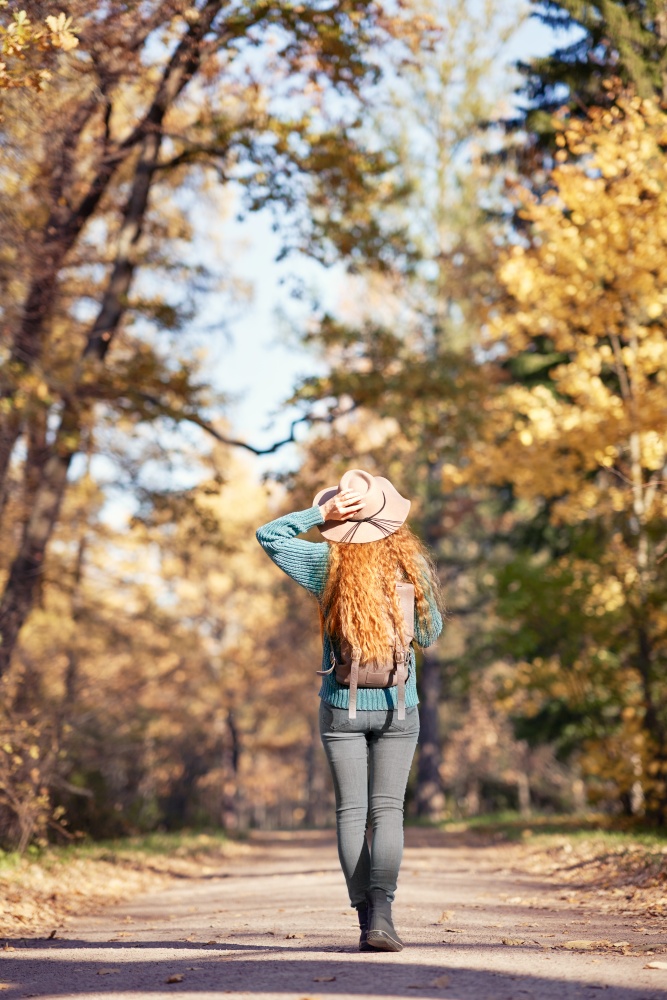 A young red-haired woman walks through the autumn park. A girl in a beige hat looks at the autumn forest