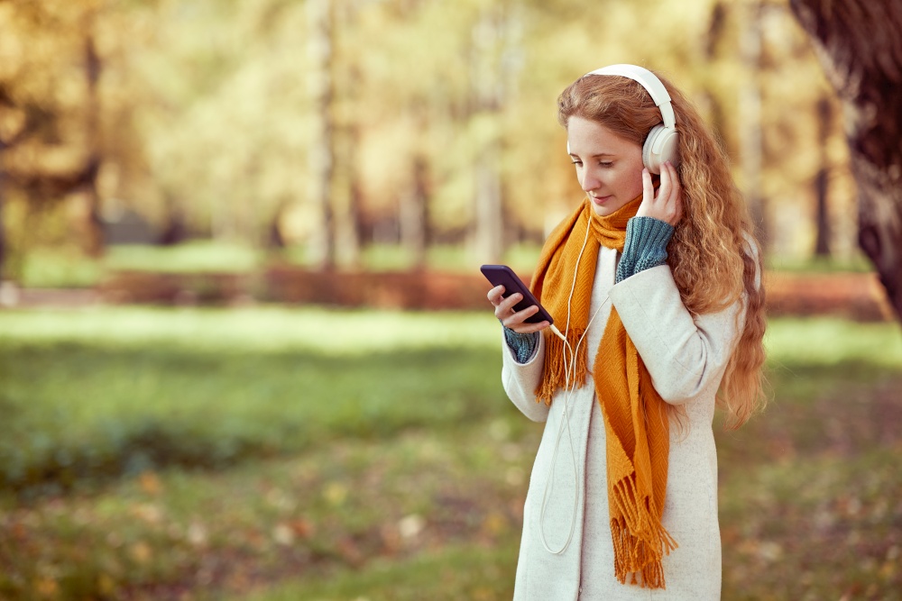 A young red-haired woman in a white coat walks in an autumn park. A girl listens to music in the autumn forest