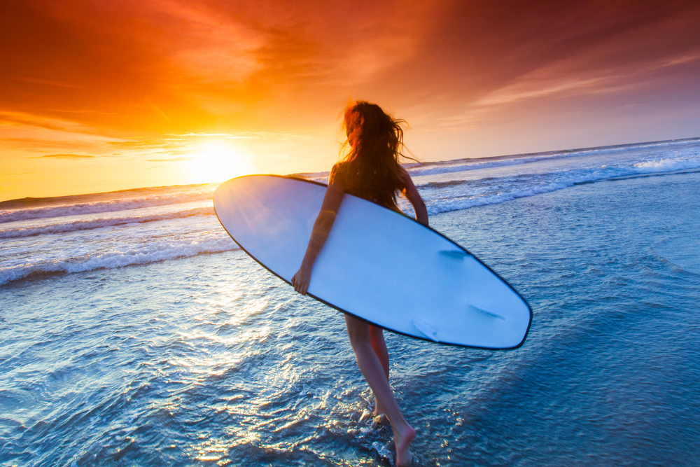 A beautiful young slim sporty woman in bikini with a surfboard is standing at ocean beach at sunset. Woman with surfboard