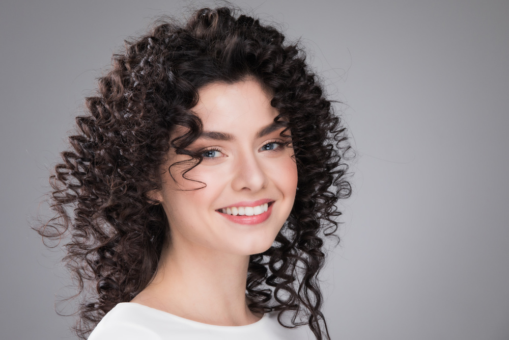 Studio portrait of beautiful smiling woman with curly dark hair on gray background. Woman with curly hair