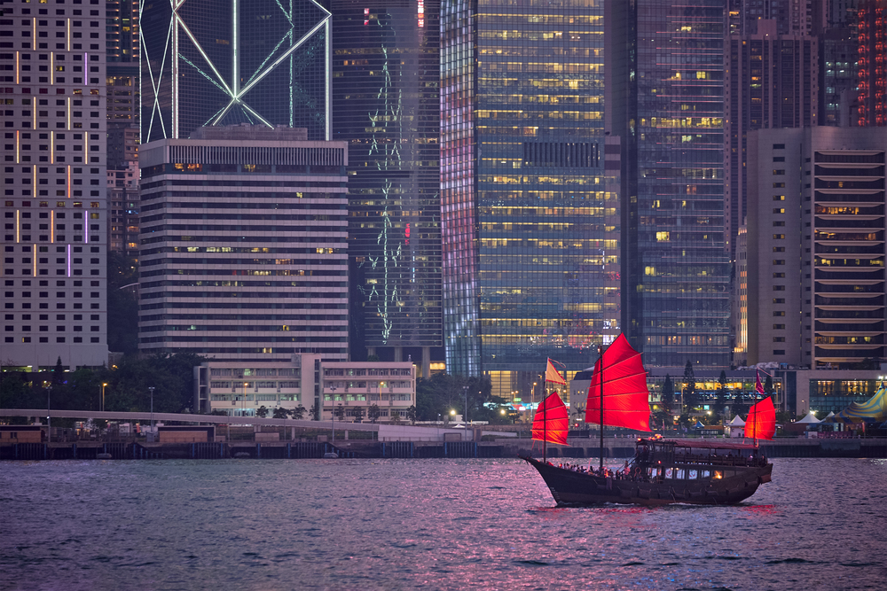 Tourist  junk boat ferry with red sails and Hong Kong skyline cityscape downtown skyscrapers over Victoria Harbour in the evening. Hong Kong, China. Hong Kong skyline. Hong Kong, China